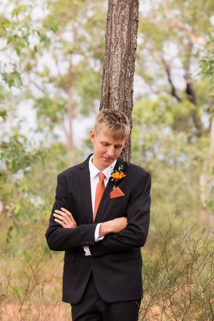 Person wearing black suit with orange tie stands thoughtfully against a tree in outdoor setting with natural lighting.