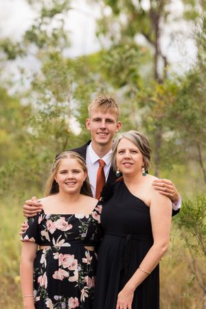 A group of three people pose together outdoors wearing formal attire with trees and greenery in the background.