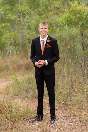A figure in a formal black suit and orange tie stands outdoors against a natural woodland background.