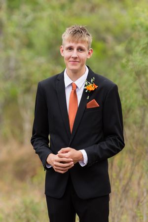 A groomsman in a black suit with orange tie smiles while standing outdoors in a blurred natural setting.