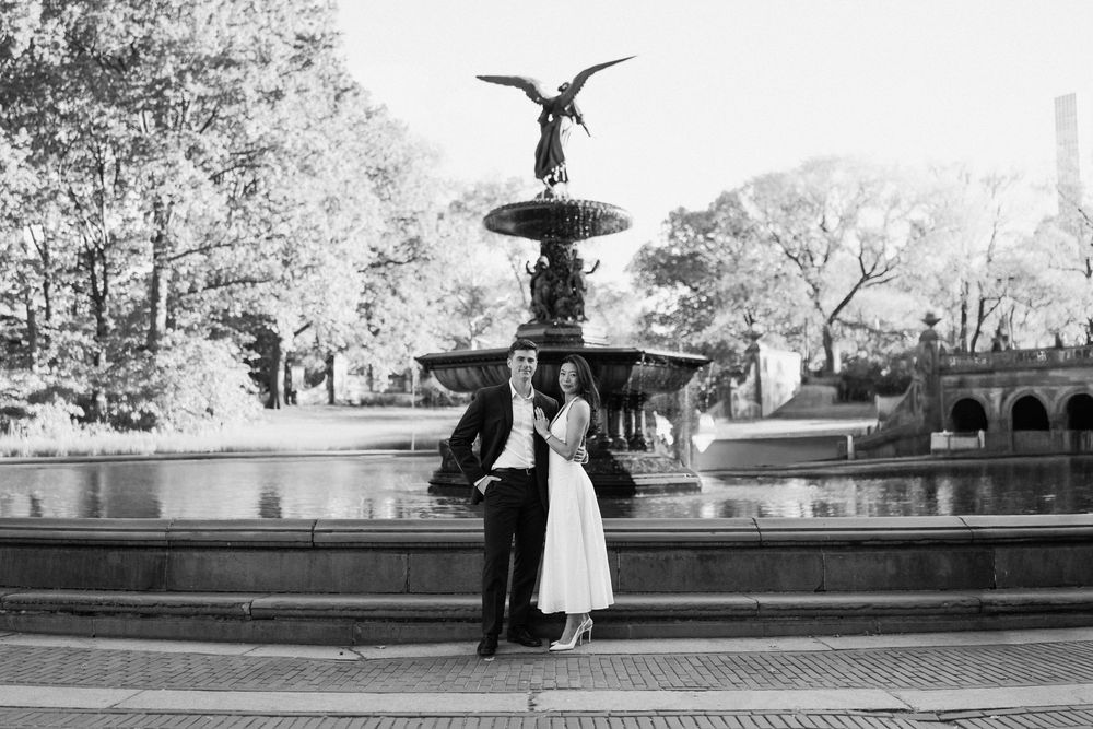 A couple poses together near a fountain in an urban park setting.