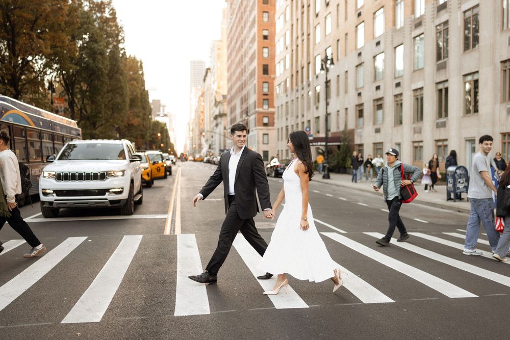 A couple crosses an urban street with city buildings in the background.