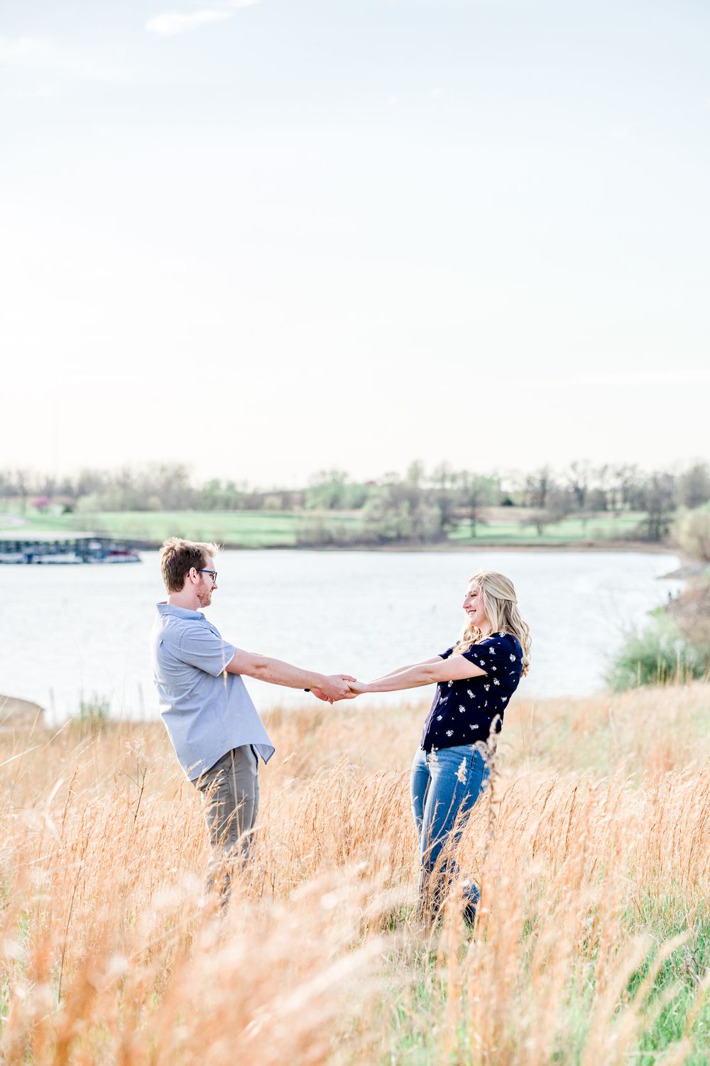 Couple holding hands and spinning in tall grass field by a lake during sunset engagement photoshoot by Stephanie Coulter Photography