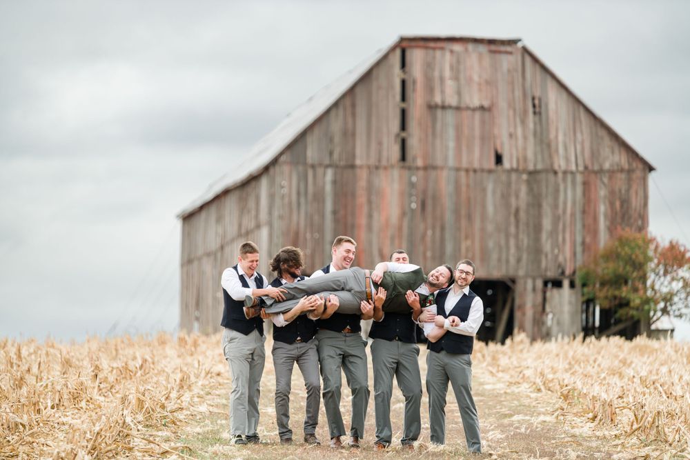 Groomsmen pose playfully while lifting groom in front of rustic barn by Stephanie Coulter Photography
