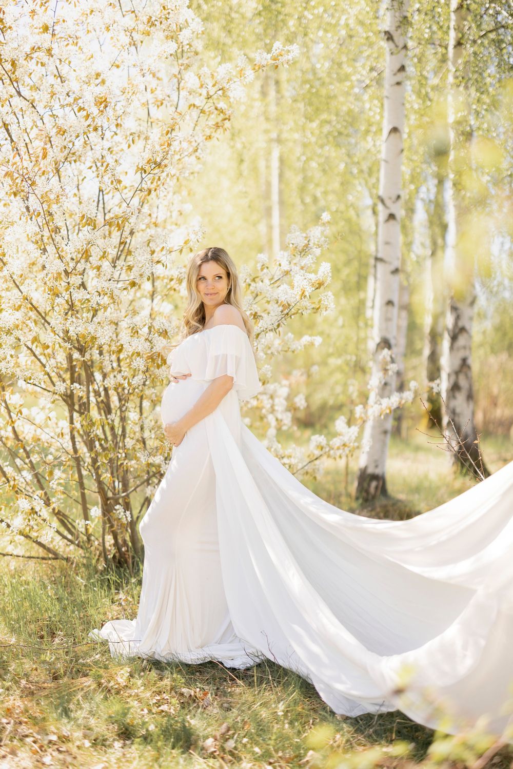 Ethereal off-shoulder white gown flows gracefully in a sunlit birch forest with blooming spring flowers.