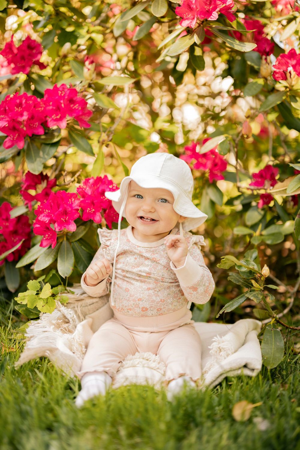 A small child sits on a blanket in a garden with vibrant pink rhododendron flowers blooming in the background.