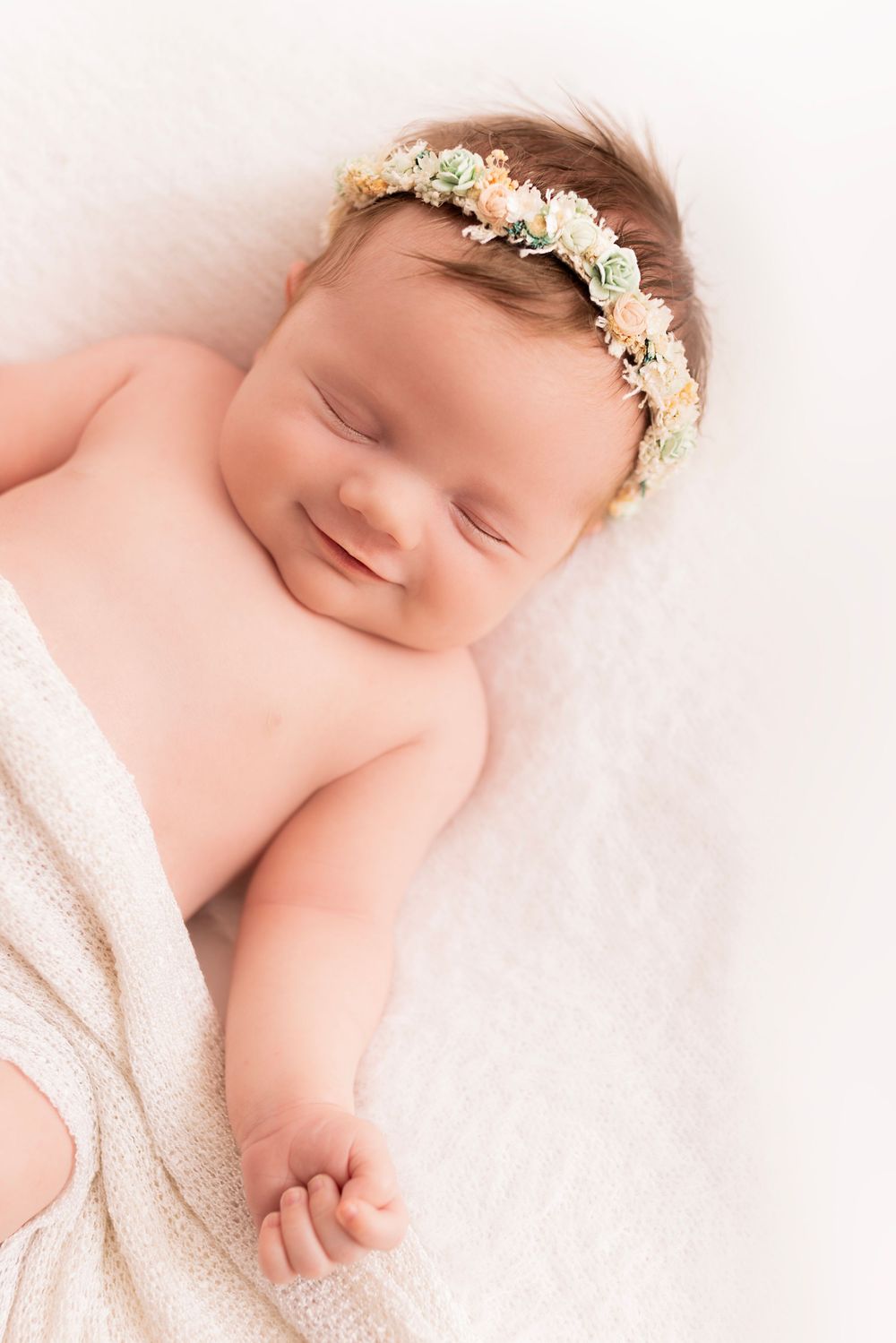 Sleeping newborn wearing delicate white floral crown headband lies peacefully wrapped in cream blanket.