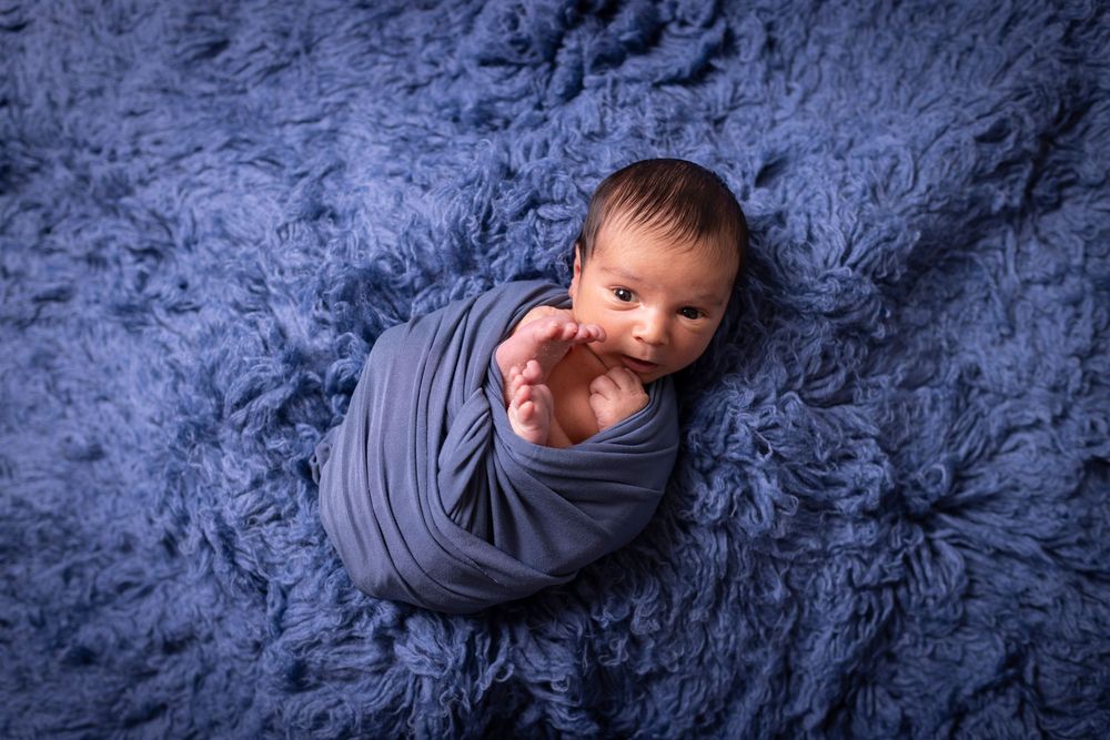 A newborn baby swaddled in gray fabric lies on a textured blue blanket during a professional photo session.