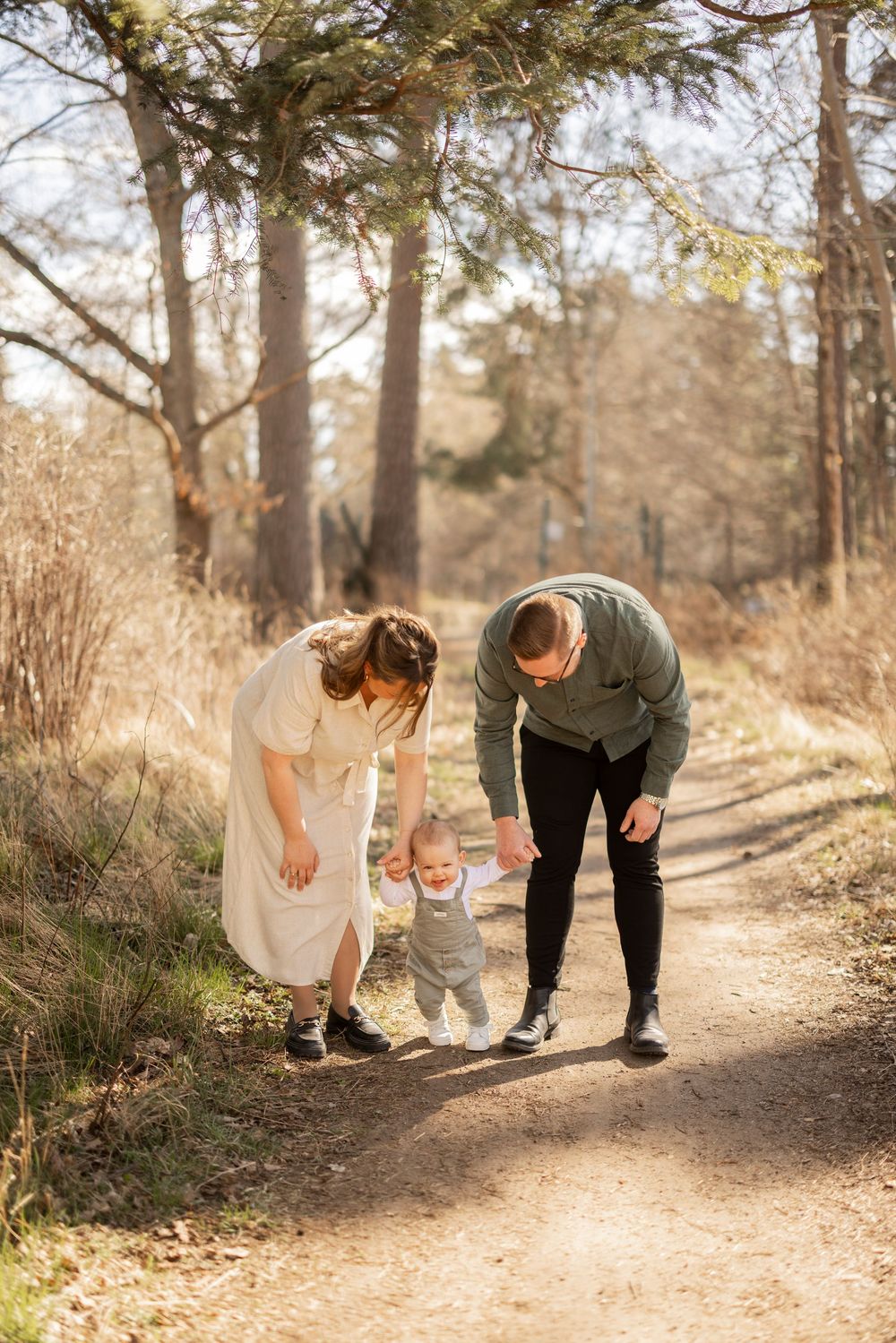 A young toddler takes supported steps on a sunlit forest trail while two adults assist on a beautiful autumn day.