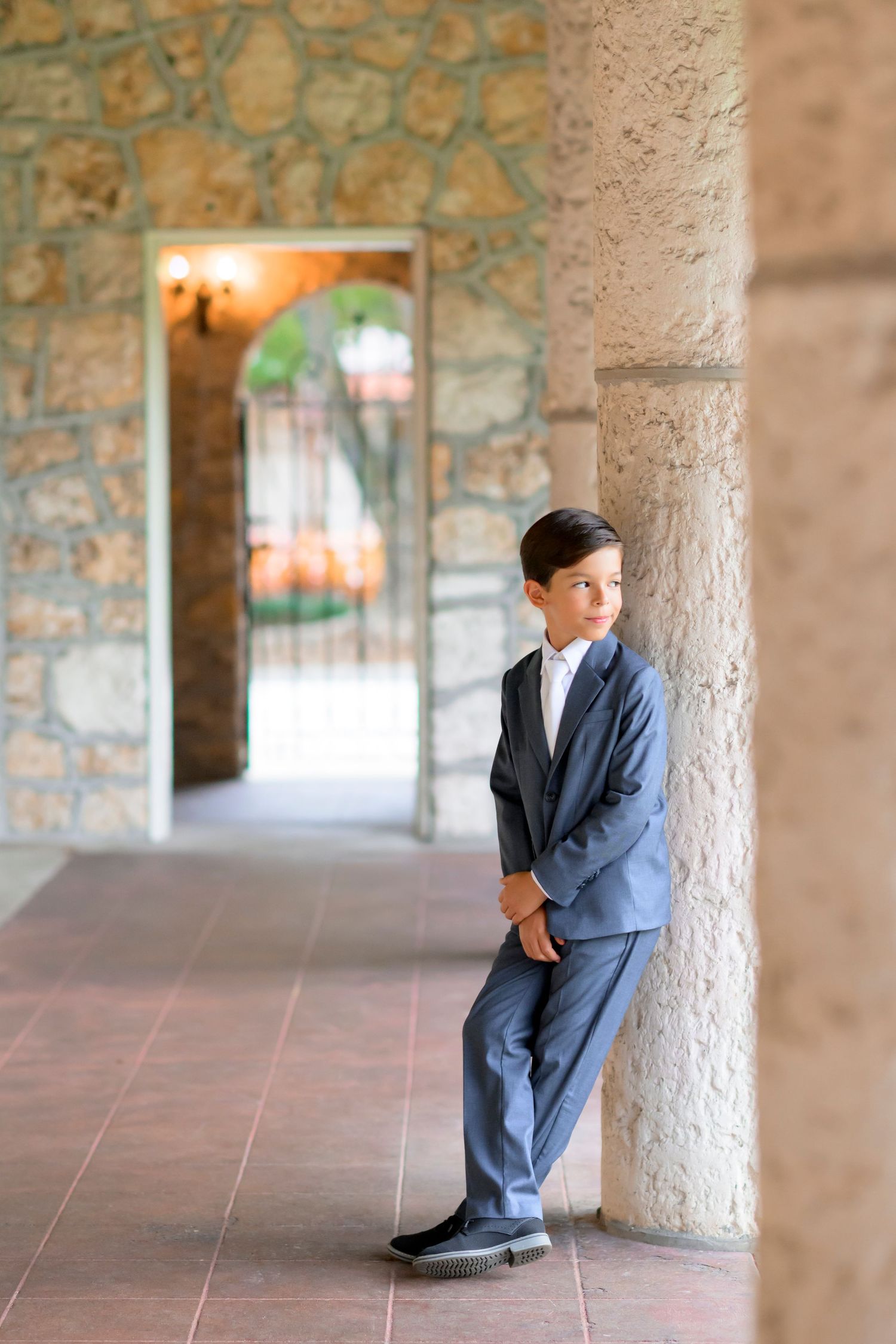 A young person in a navy blue suit leans against a stone pillar in an elegant covered walkway with arched openings.