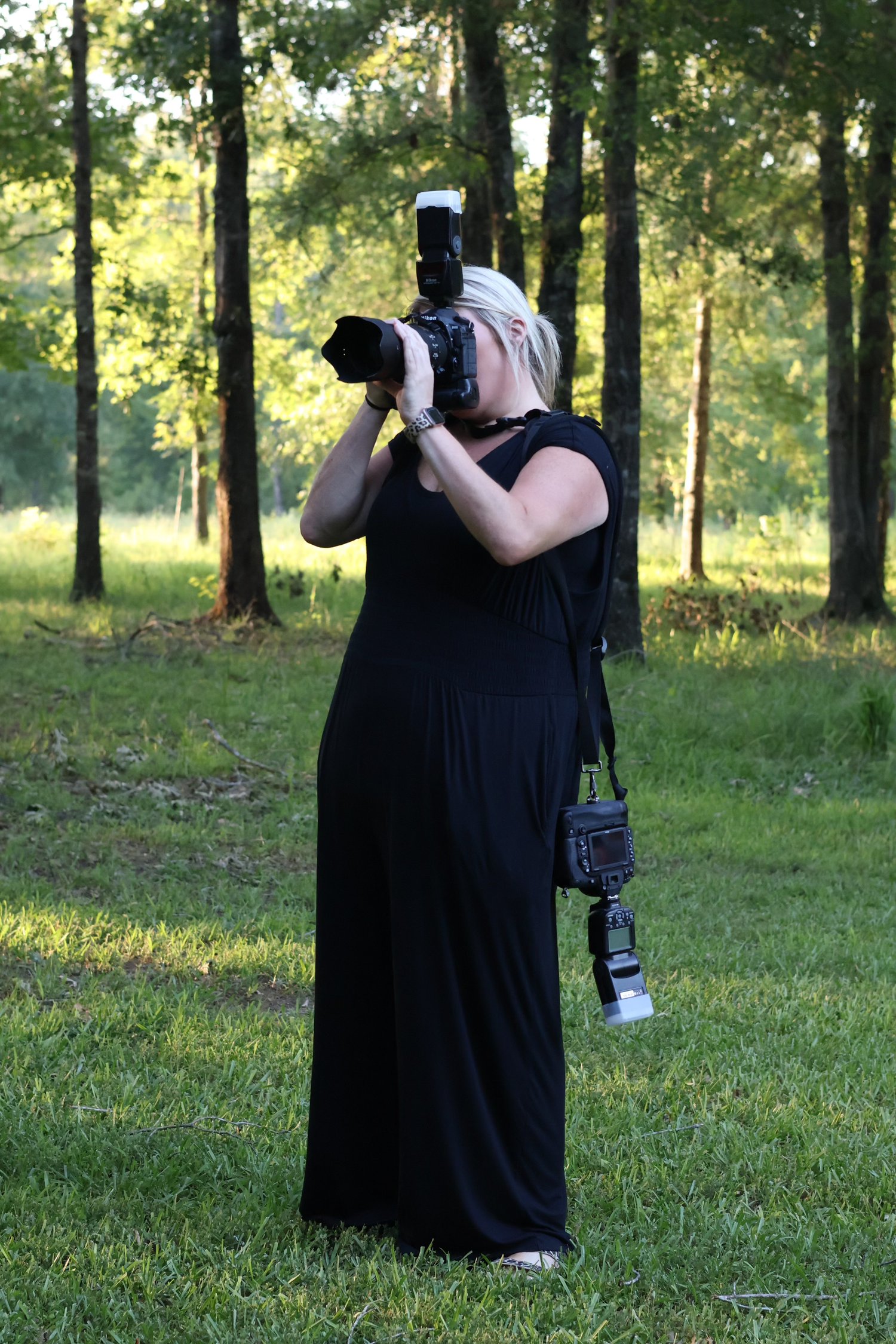 Professional photographer in black dress takes photos in sunlit wooded setting.