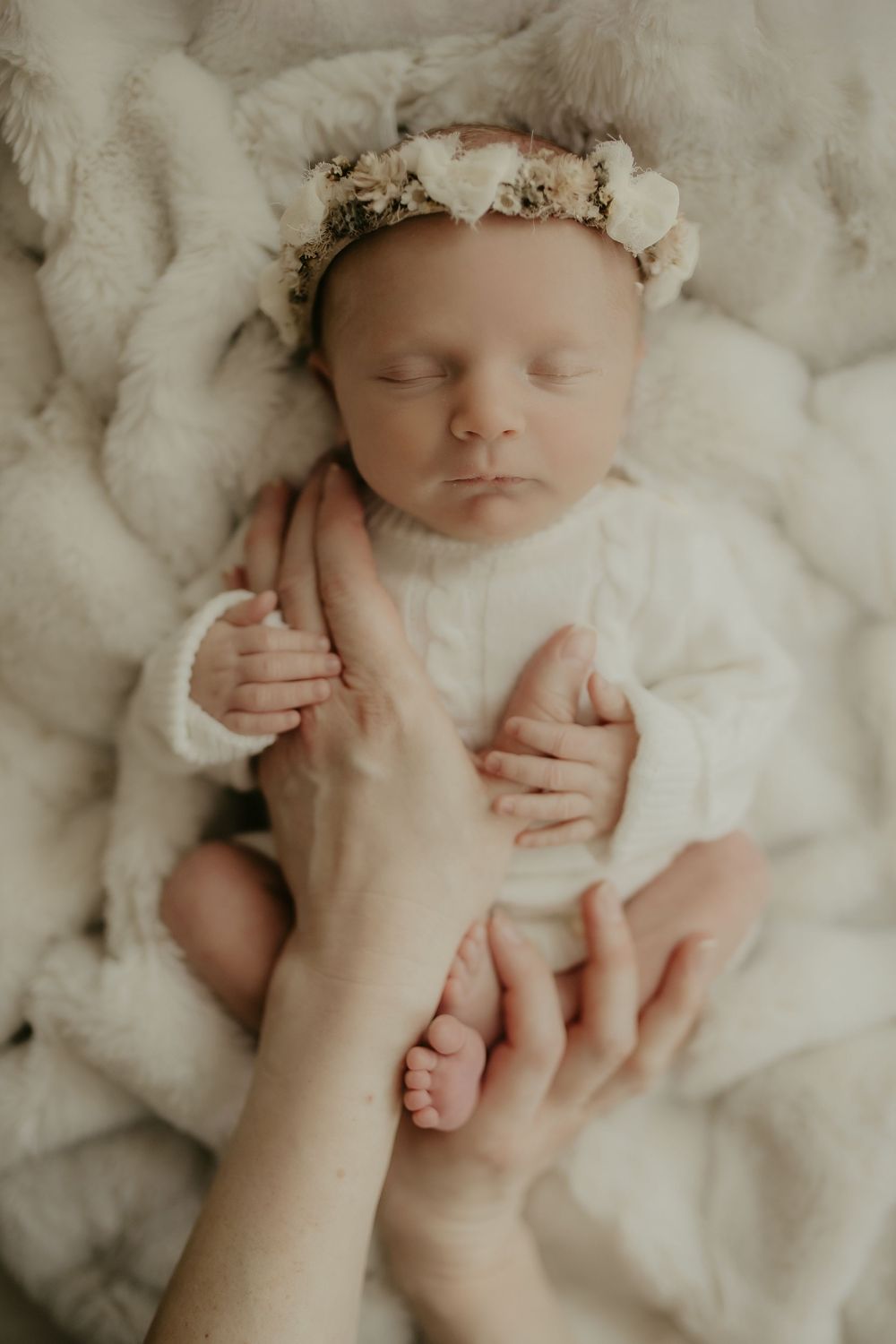 Newborn baby wearing delicate floral crown rests peacefully on soft white blanket in professional photo series.
