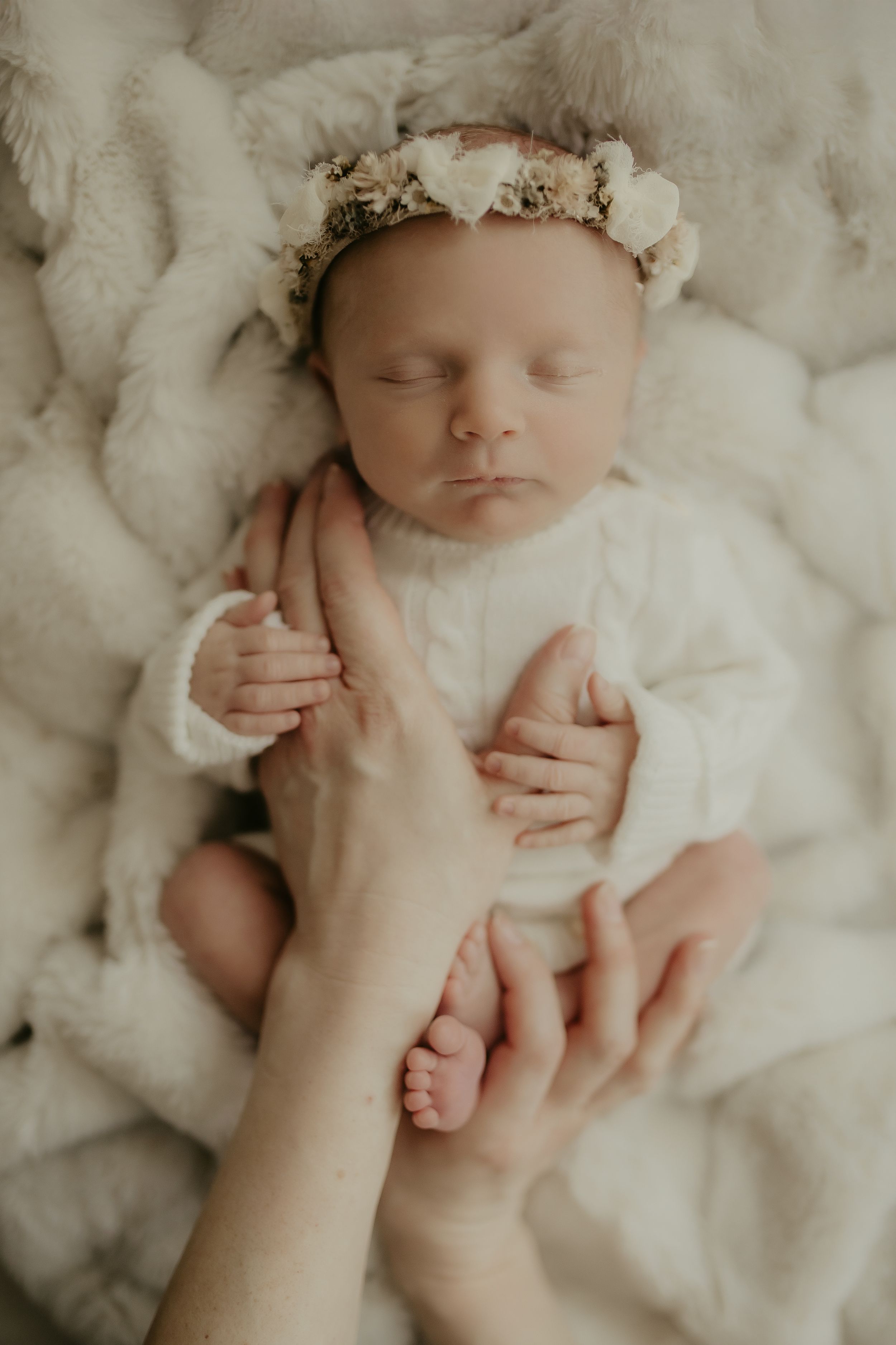 Newborn baby wearing delicate floral crown rests peacefully on soft white blanket in professional photo series.