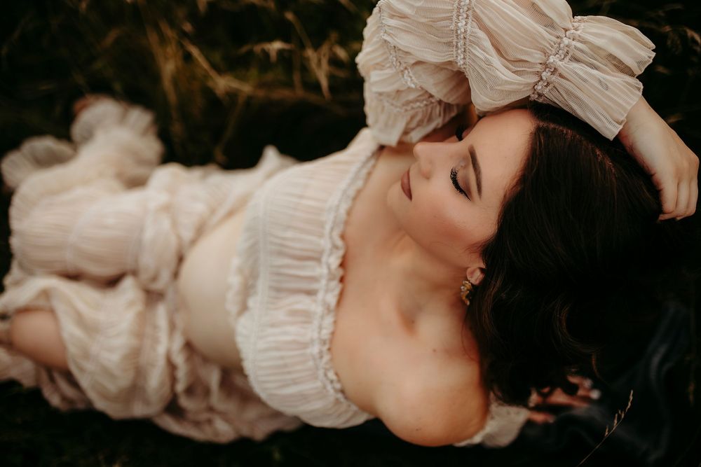 A woman in a white ruffled dress with a bow lies gracefully among dark green foliage.