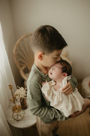 A tender moment between siblings as an older child in gray gently embraces and kisses a baby in white lace clothing.