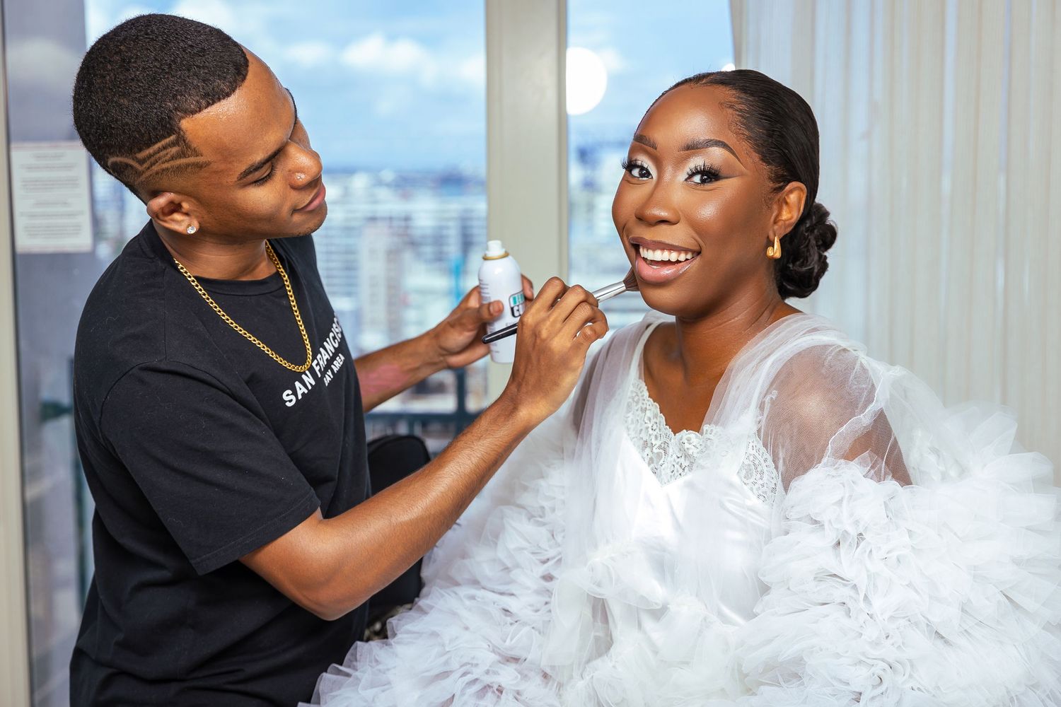 Makeup artist applies cosmetics to smiling woman in white feathered outfit.