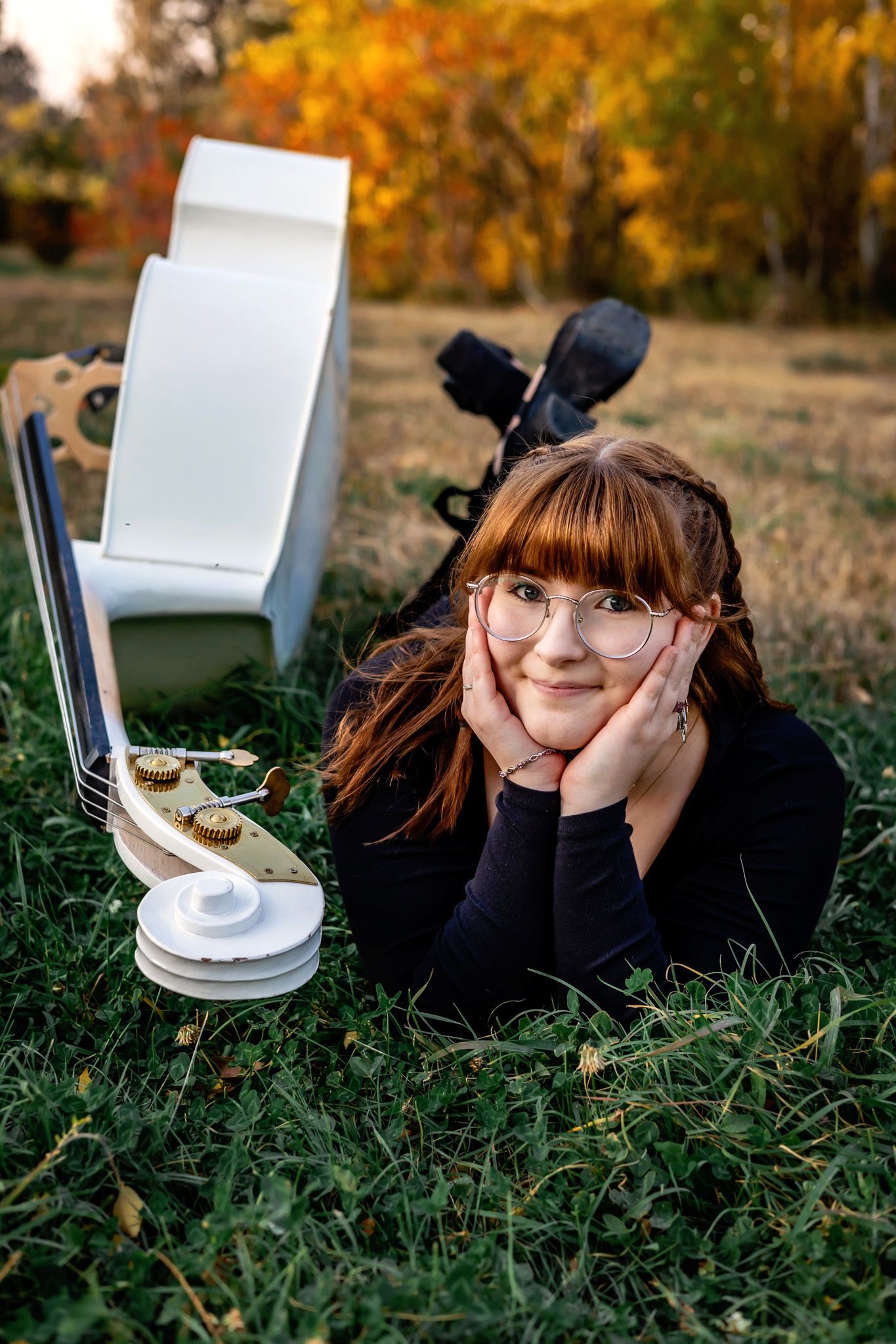 A student lies on grass studying with a bass during autumn while wearing glasses and a black sweater.