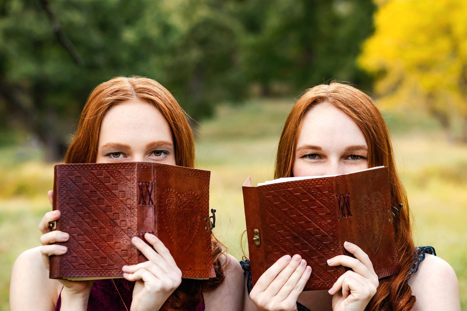 Twin sisters hold matching leather-bound books up to cover the bottom half of their faces.