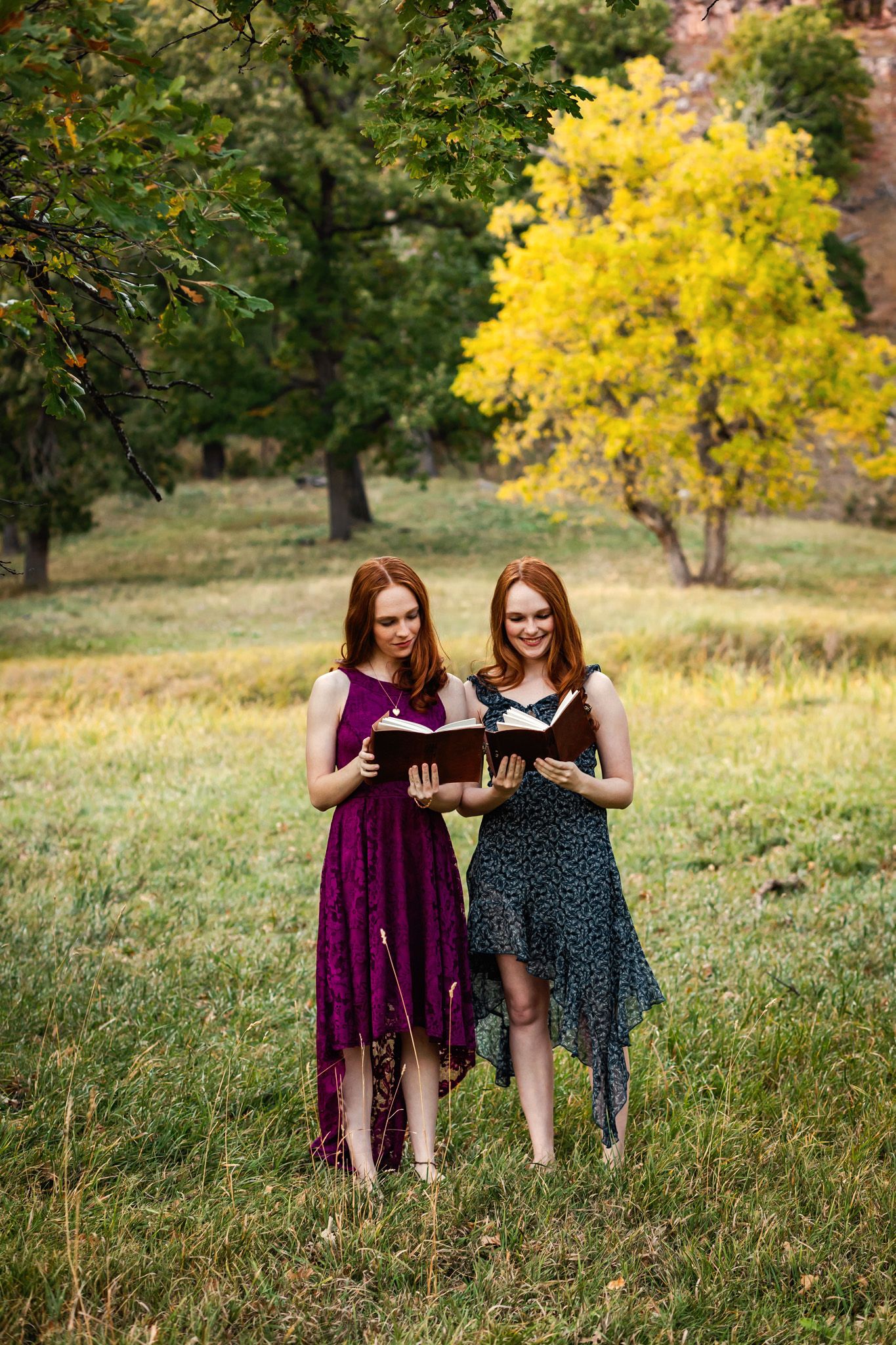 Twins in dresses reading books together while standing in a grassy field with autumn trees in the background.