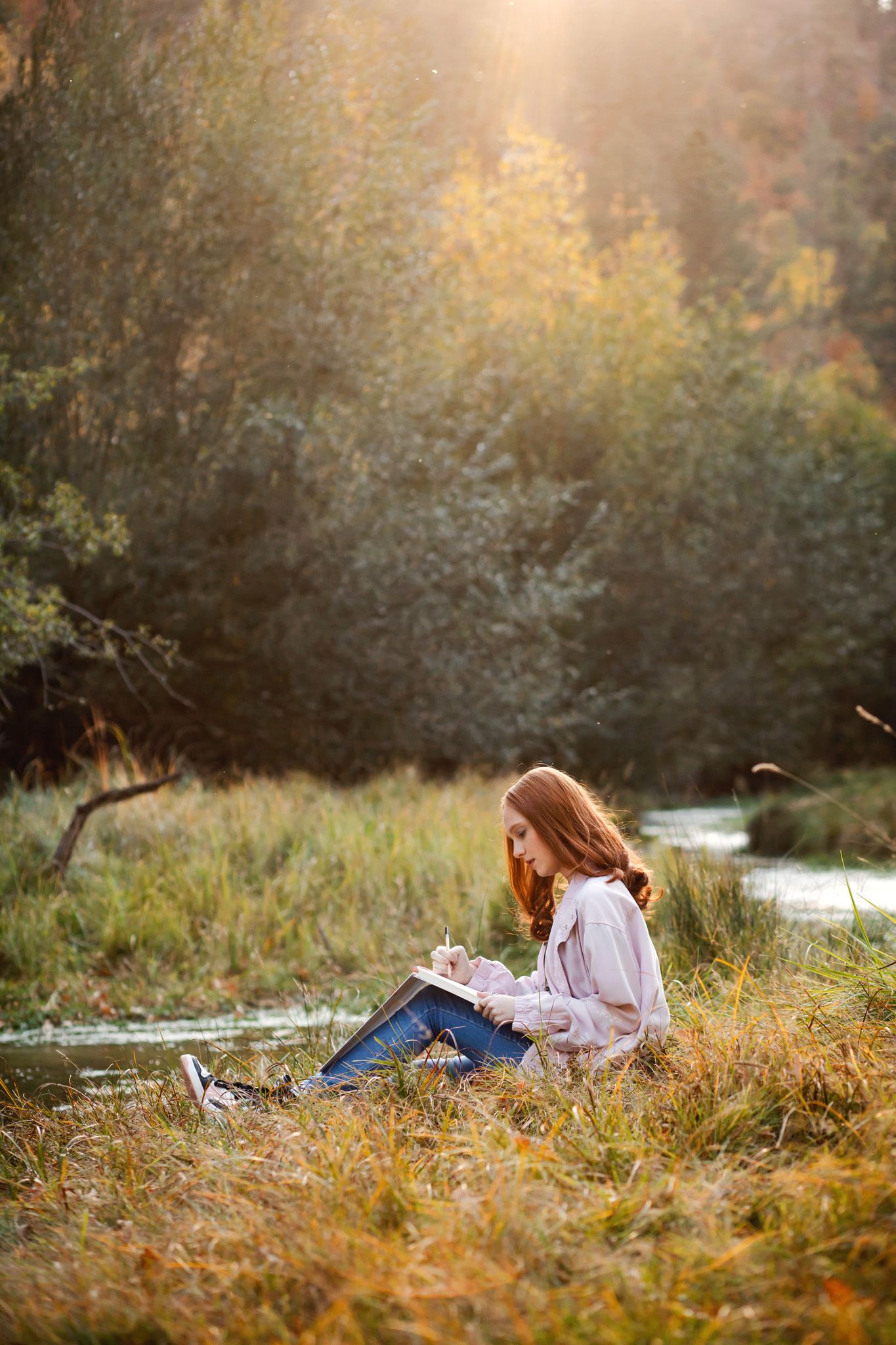 Girl sits writing in a journal by a creek during golden hour with fall foliage in background.