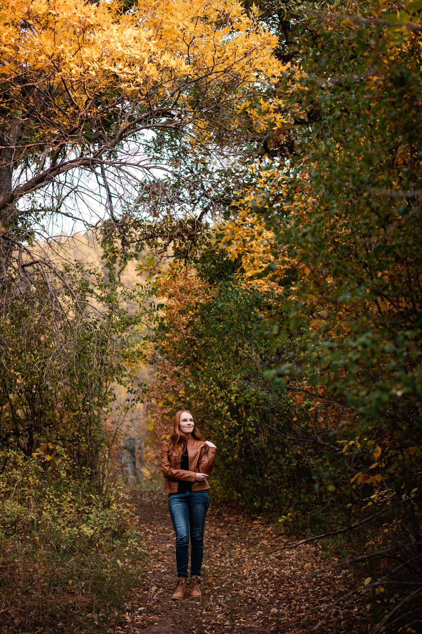 A girl walks along a leaf-covered forest trail surrounded by trees with vibrant golden autumn foliage.