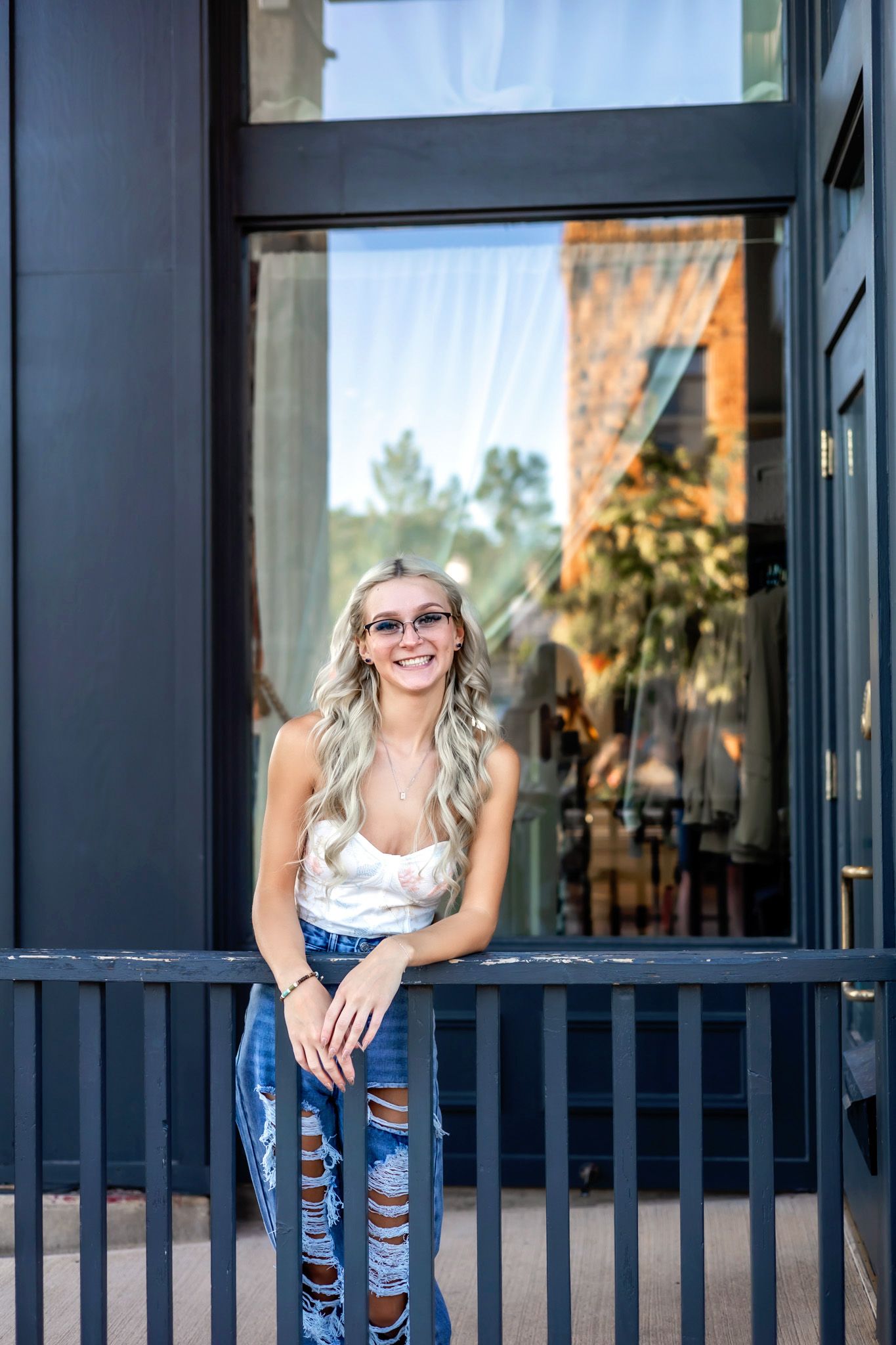 Casual fashion portrait of a smiling woman in ripped jeans and white top leaning against dark railing outside storefront.