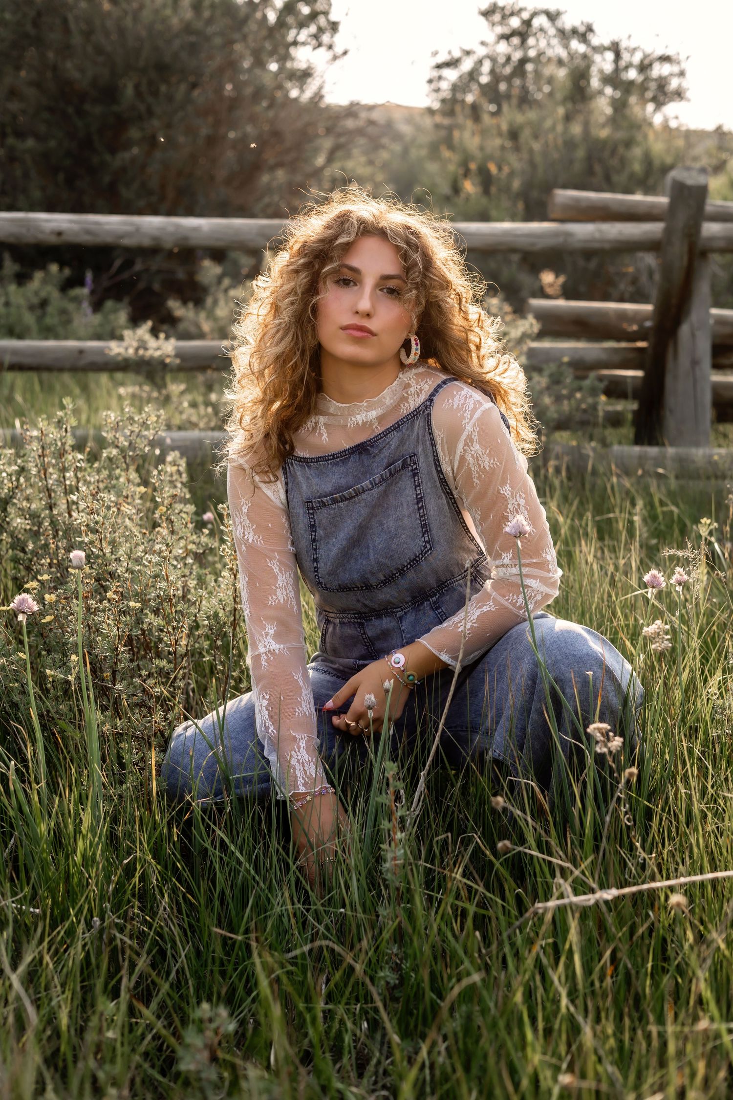 A girl in denim overalls and lace top poses in a sunlit meadow with wildflowers near a rustic wooden fence.
