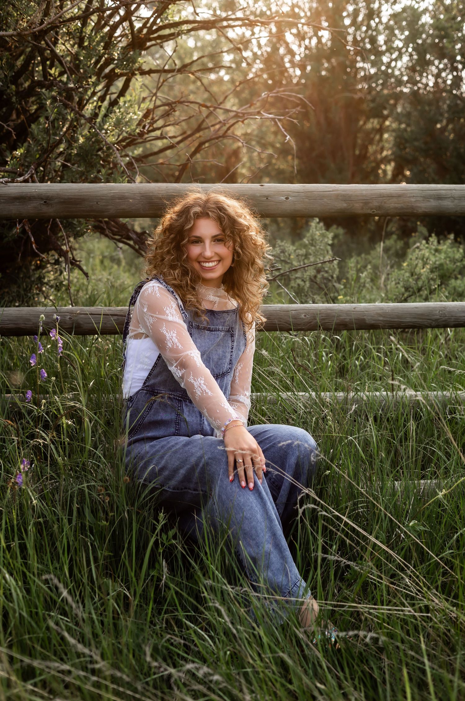 Girl wearing denim overalls and lace top sits by rustic wooden fence at sunset in tall grass field.