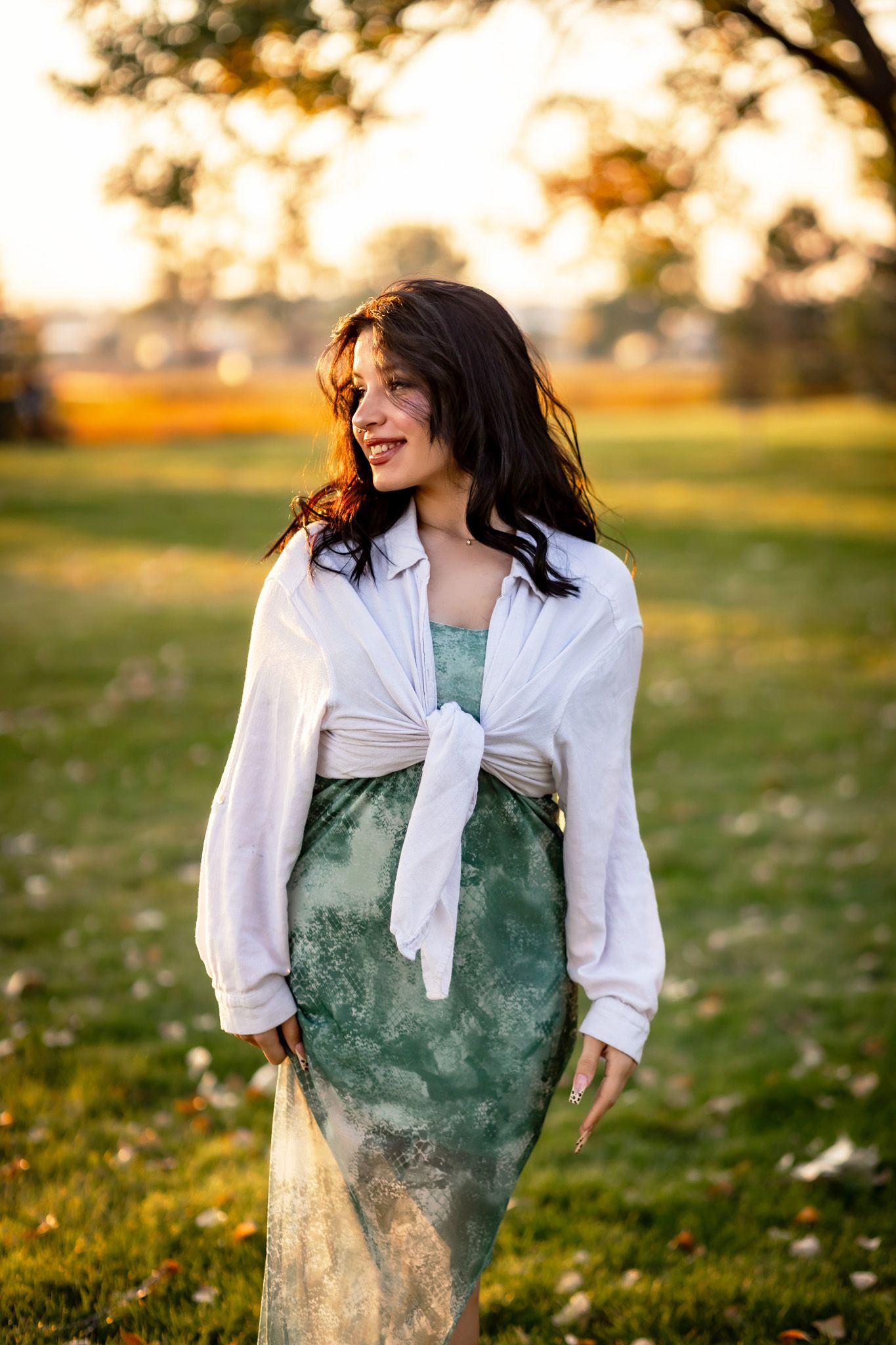 A woman in a white blouse and green patterned dress stands in a sunny field during golden hour.