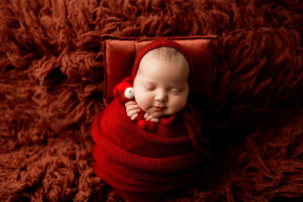 A sleeping baby wrapped in a red knit blanket lies in a small box against a deep burgundy textured background.