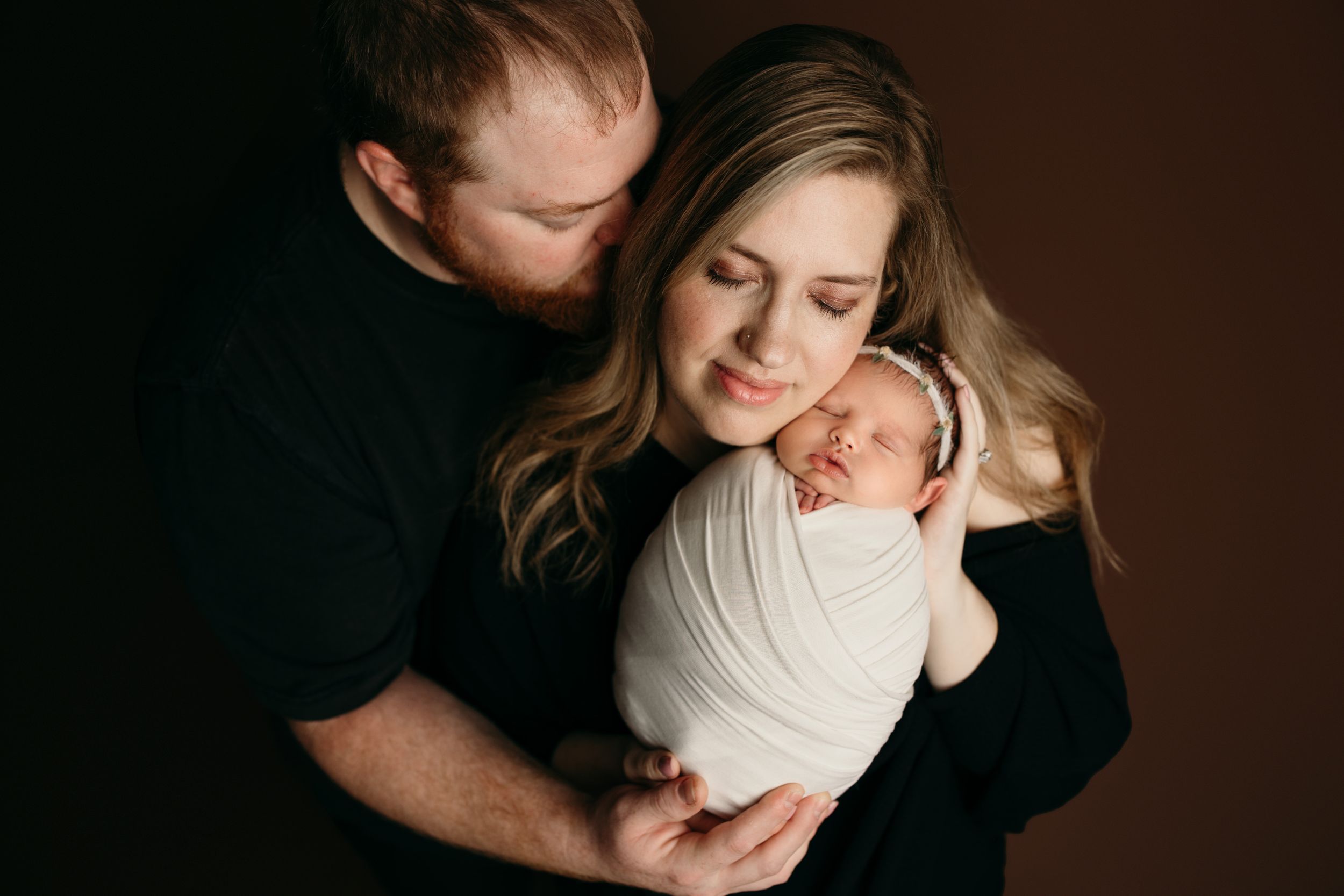 A newborn baby wrapped in white swaddle is held close by parents during an intimate studio portrait session.