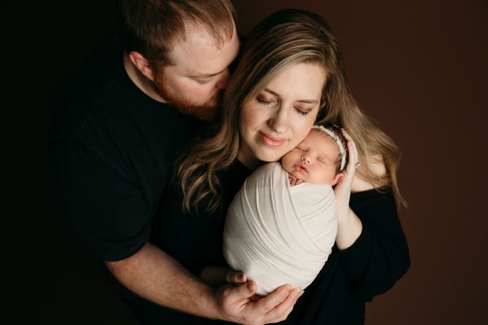 A newborn baby wrapped in white swaddle is held close by parents during an intimate studio portrait session.
