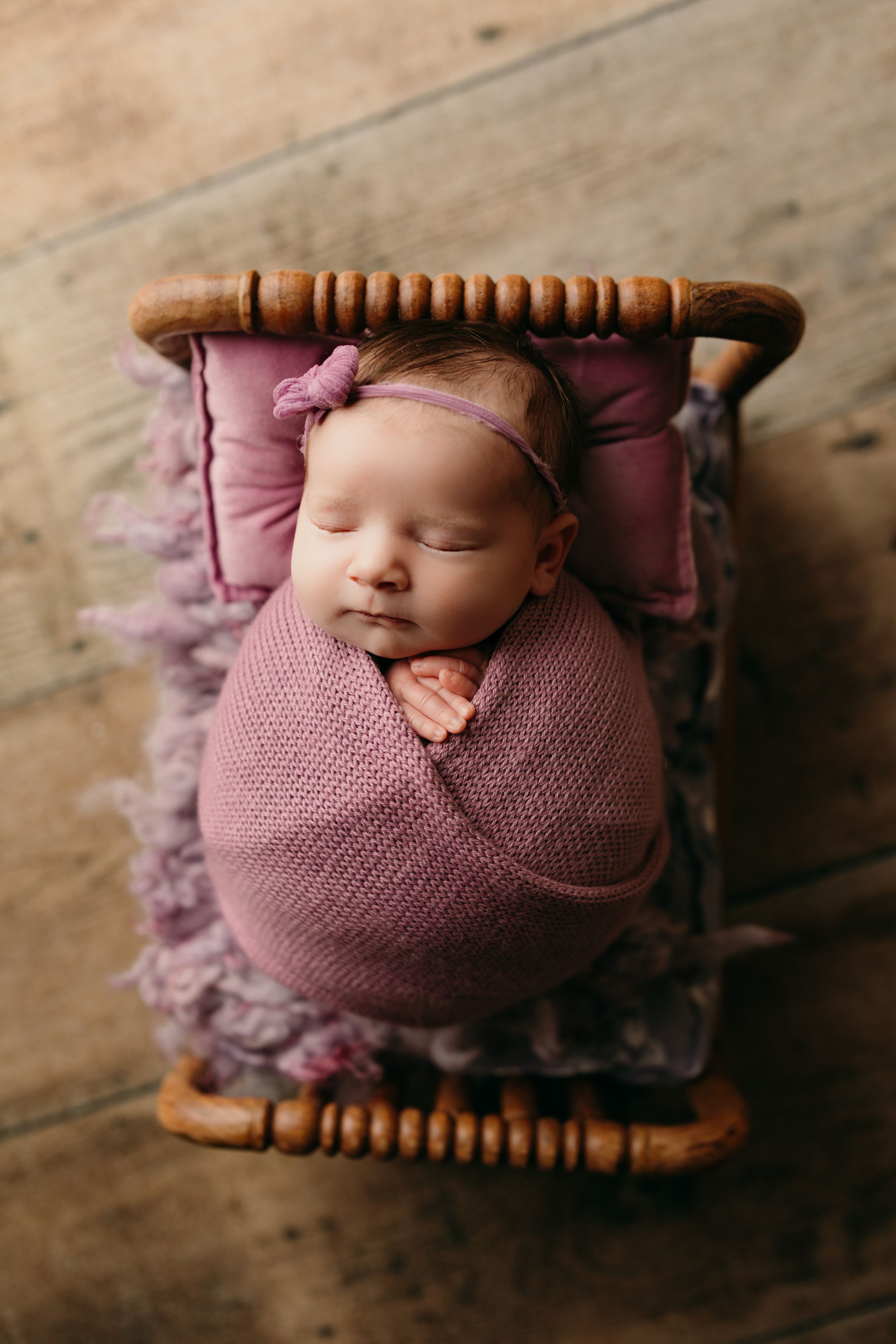 Newborn photography series showing baby wrapped in mauve knit blanket on wooden spool prop against rustic floor.