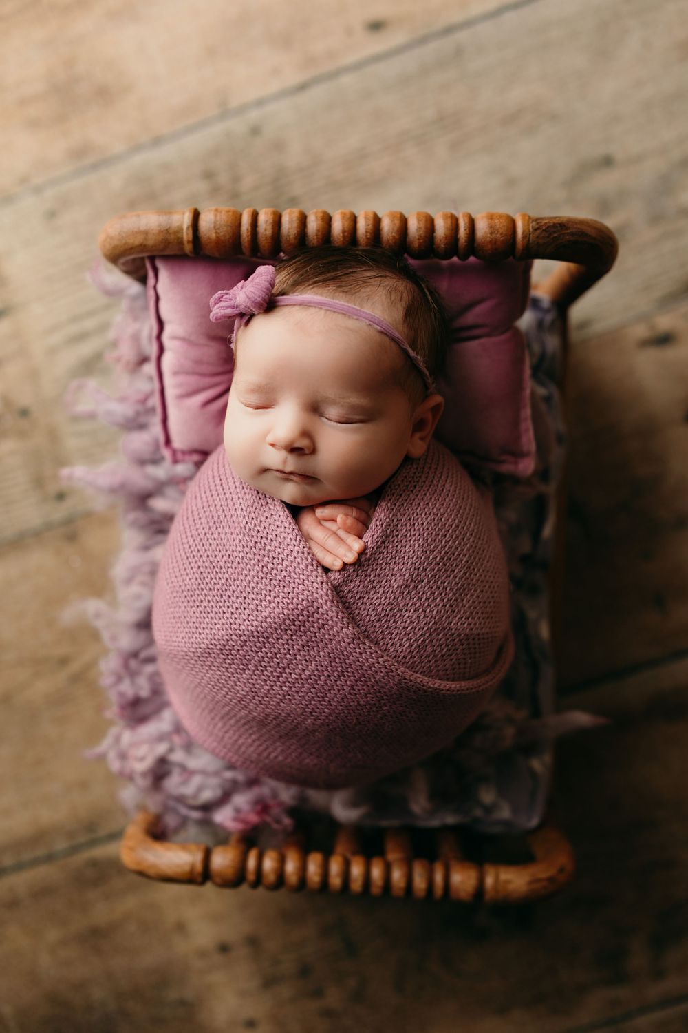 Newborn photography series showing baby wrapped in mauve knit blanket on wooden spool prop against rustic floor.