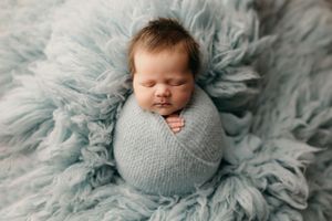 Sleeping newborn wrapped in soft gray blanket on light blue textured backdrop.