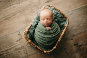 Newborn sleeping peacefully in woven basket with sage green wrap.