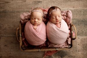 Twin newborns swaddled in pink wraps sleeping side by side in wooden basket.