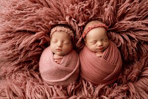 Sleeping newborn twins nestled together on soft pink fluffy backdrop.