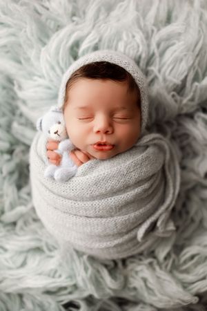 Baby wrapped in soft grey swaddle blanket on fluffy textured background.