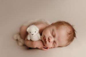 A sleeping newborn baby lies peacefully next to a small white plush lamb toy in a neutral-toned setting.