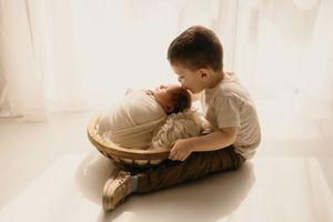 Tender moment between siblings as older child leans over to kiss baby in woven basket by window.