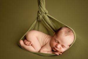 A newborn baby sleeps peacefully while suspended in a soft green fabric sling against an olive background.