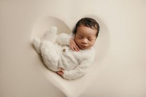 A sleeping newborn baby wrapped in soft white blankets lies peacefully against a cream-colored backdrop.