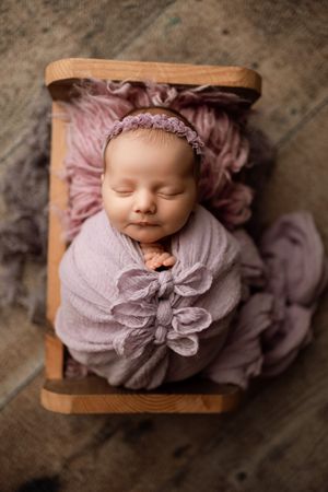 Sleeping newborn in wooden bed with mauve wrapping and delicate headband.
