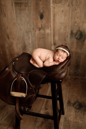 Sleeping newborn posed on brown leather saddle against rustic wooden background.