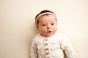 Sweet newborn in white knit outfit with delicate headband on cream background.