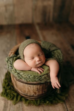 A sleeping newborn baby nestled in a rustic wooden bowl with olive green fabric and natural textures.