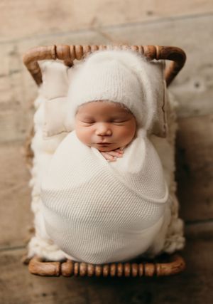A sleeping baby wrapped in white knit blanket rests peacefully on a rustic wooden surface in a creative newborn photo.