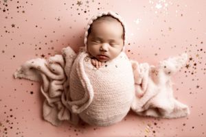 Sleeping newborn wrapped in soft pink swaddle blanket lying on pink background with scattered sparkle effects.