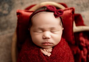 Newborn sleeping on burgundy fabric with matching headband against rustic backdrop.